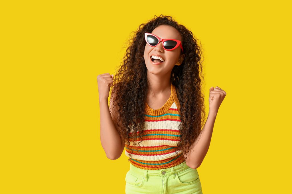 Mulher alegre com roupas coloridas e óculos vermelhos em fundo amarelo vibrante, representando a paleta de verão.