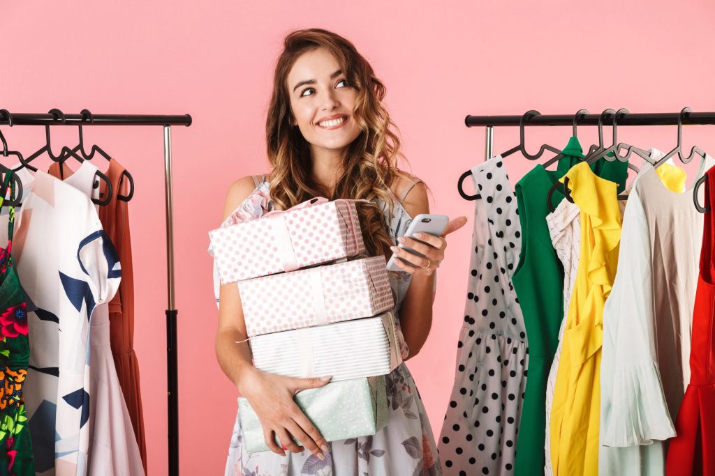 Mulher segurando caixas de compras ao lado de araras com roupas estampadas, ilustrando o apelo visual na moda.